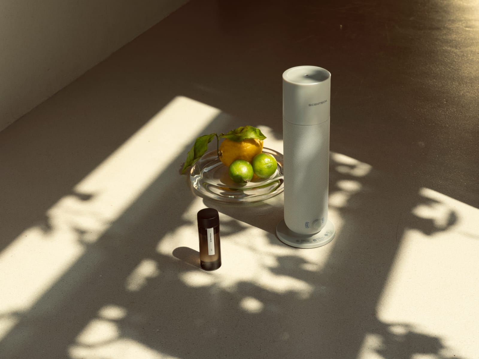 Smart Aroma Diffuser 2 met een geurflesje ernaast en een schaal limoenen, in gevlekt zonlicht op tafel